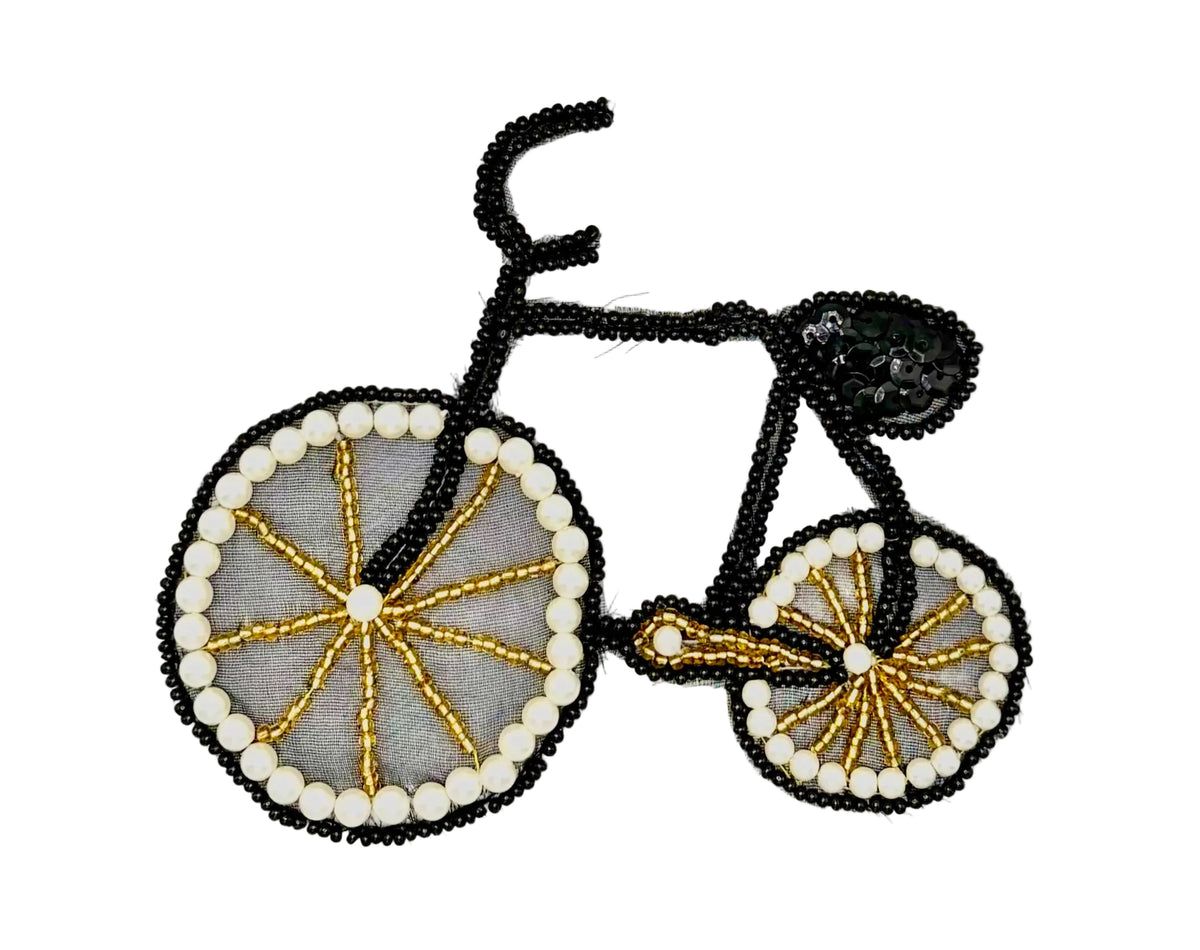 A bicycle-shaped decorative patch or applique made from black sequins, black seed beads, white pearl-like beads, and gold-toned metallic beads on a light gray background.