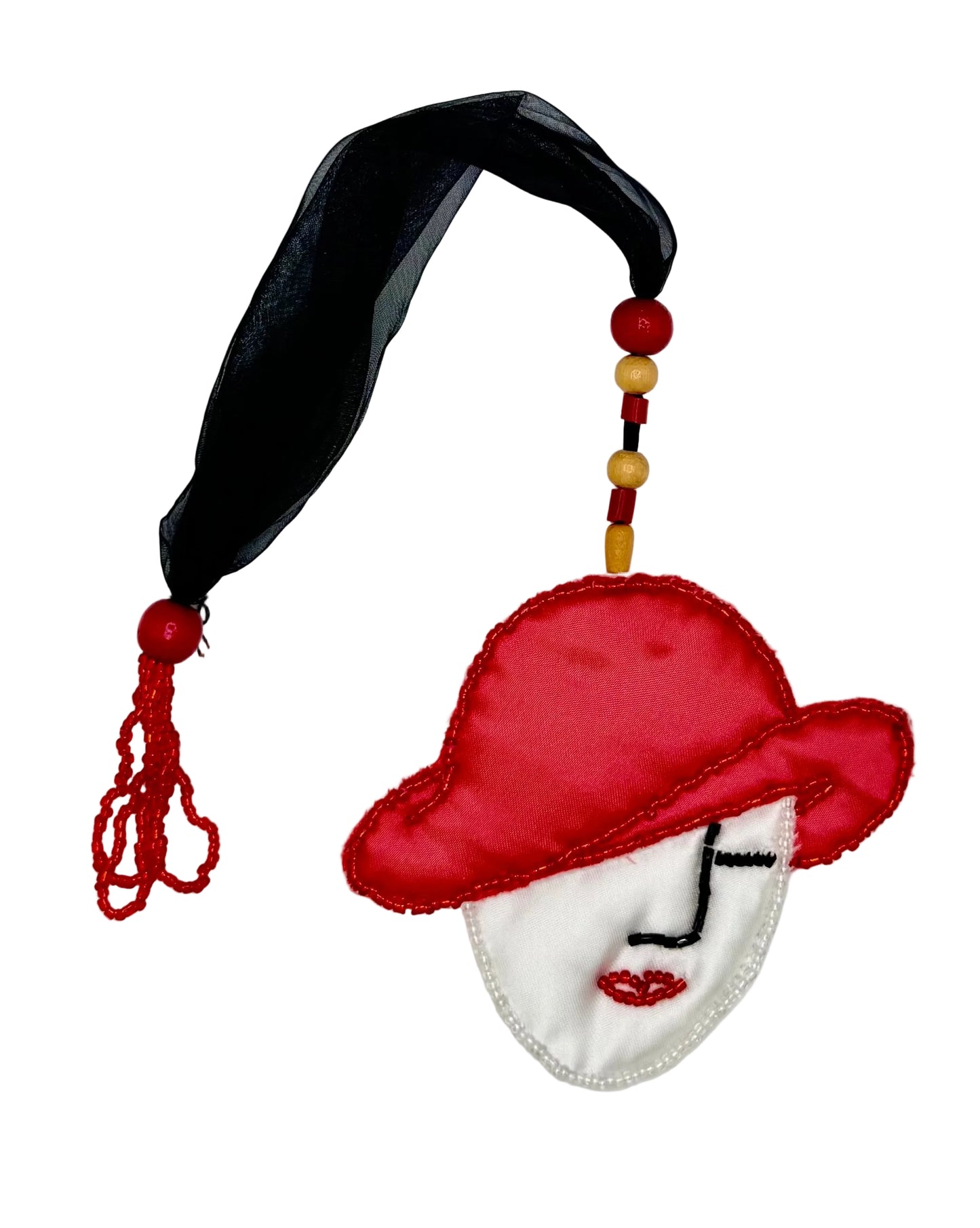 A decorative fabric ornament featuring a stylized face with a red hat, white face, black beaded eyes and nose, red lips, a black ribbon loop, and red and yellow beads with a tassel on a plain white background.