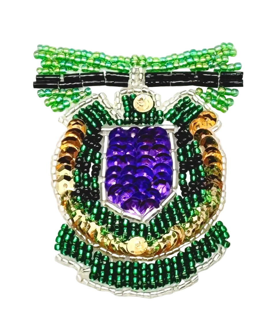 A beaded decorative crest featuring a purple sequin shield in the center, surrounded by green and black seed beads, gold-toned accents, and a black ribbon motif at the top, shown on a plain white background.