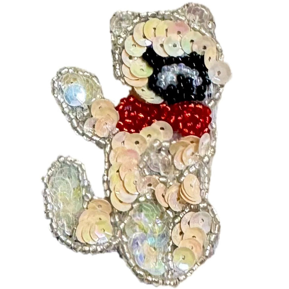 A teddy bear-shaped embellished brooch or pin made of iridescent sequins, featuring a black face, red beaded scarf, and silver-toned sequined border on a plain white background.