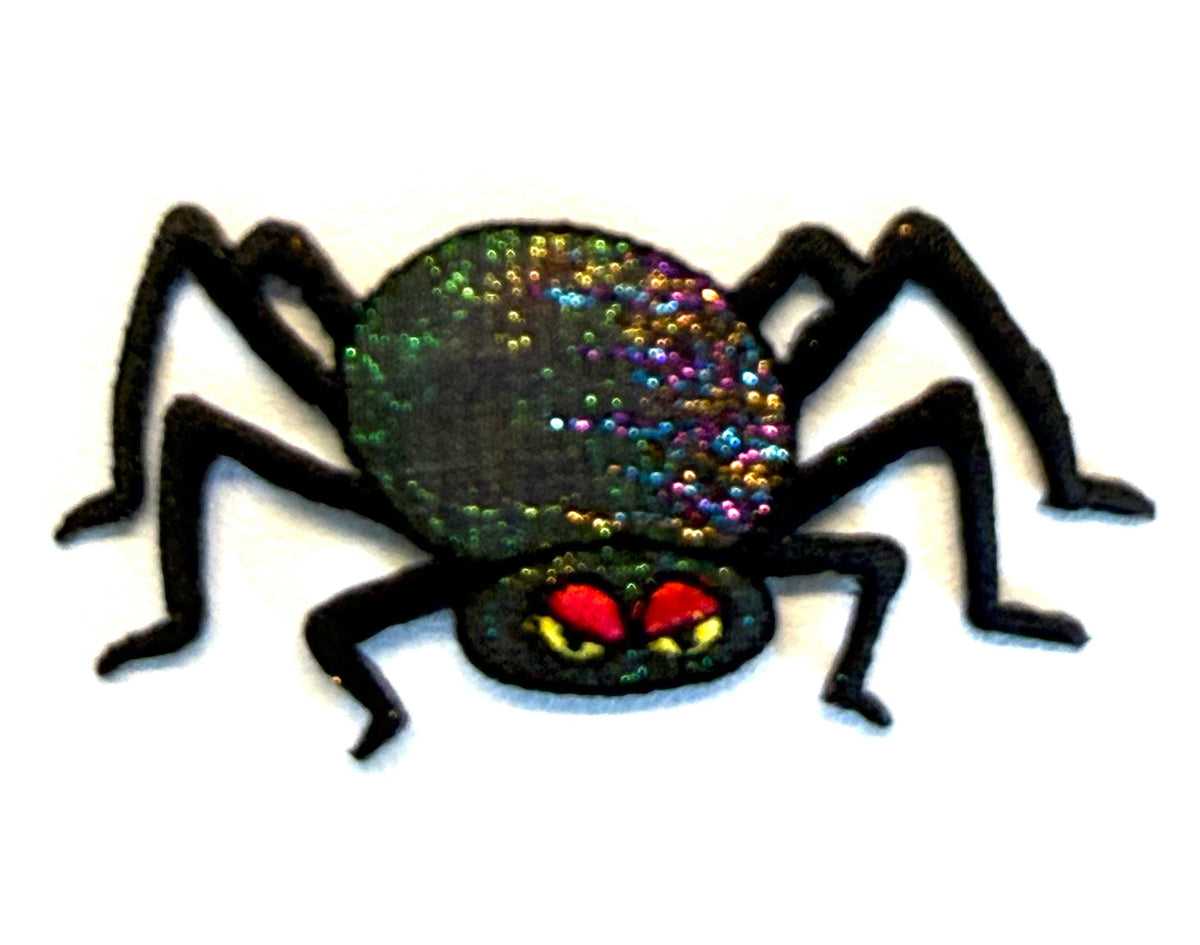 A black spider-shaped patch with a glittery, iridescent green and purple abdomen and red eyes with yellow details, shown on a plain white background.