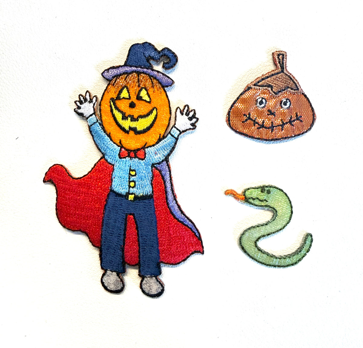 Three embroidered Halloween-themed patches on a white background: a jack-o'-lantern figure in a blue hat and red cape, a brown acorn patch, and a green snake patch.