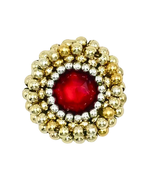 A vintage-style round patch featuring a central faceted red gemstone surrounded by a halo of textured gold and silver metallic beads, set against a plain white background.