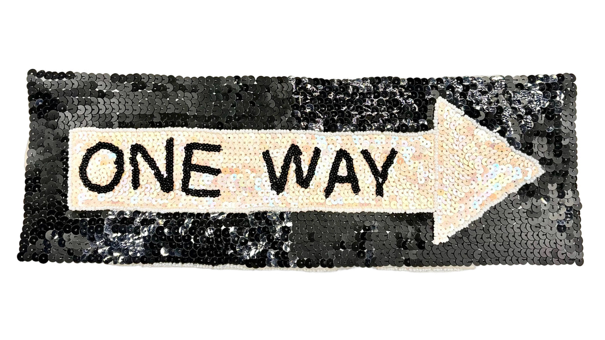 A rectangular sequin patch shaped like an arrow pointing right, with a cream center and the words "ONE WAY" in black lettering, set against a black sequined background.