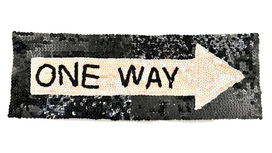 A rectangular sequin patch shaped like an arrow pointing right, with a cream center and the words "ONE WAY" in black lettering, set against a black sequined background.