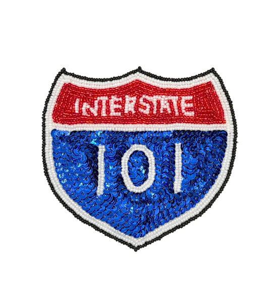A shield-shaped sequin and bead patch designed like the Interstate 101 highway sign, featuring a red top section with white 'INTERSTATE' text, a blue lower section with white '101' text, and a black and white stitched border on a plain white background.
