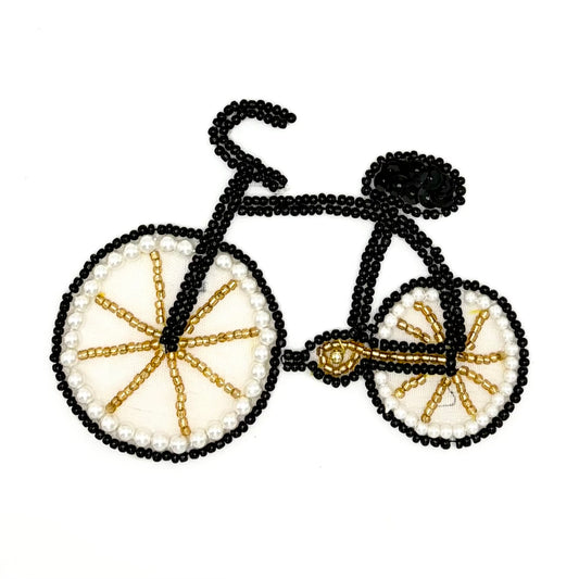A bicycle-shaped decorative patch or applique made from black seed beads, with cream circular wheels outlined in black and accented by gold seed beads, shown on a plain white background.