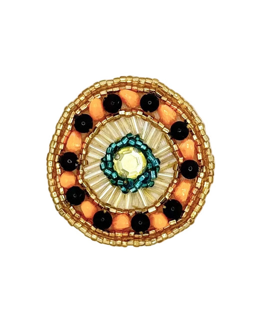 A circular beaded accent featuring a gold-toned outer border, orange and black beads, and a central turquoise and cream sunburst design with a clear faceted gem.