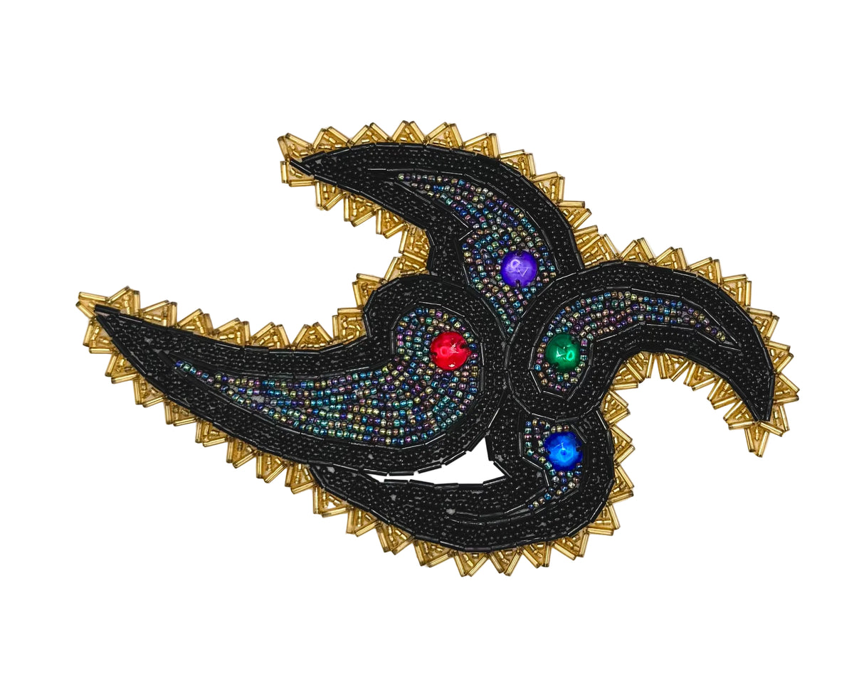 A decorative beaded patch featuring a stylized three-petal or triskelion motif in black and gold, embellished with iridescent beads and four colorful rhinestones in red, green, blue, and purple, set against a plain white background.