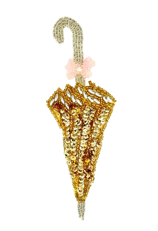 A miniature umbrella-shaped appliqué embellished with gold and silver sequins and beads, featuring a light pink beaded bow at the top and a curved silver handle, set against a plain white background.
