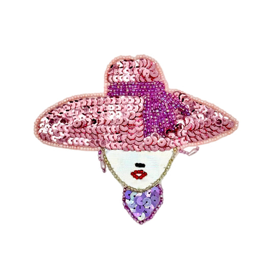 A decorative sequined patch shaped like a woman's face wearing a large pink hat shown on a plain light background.