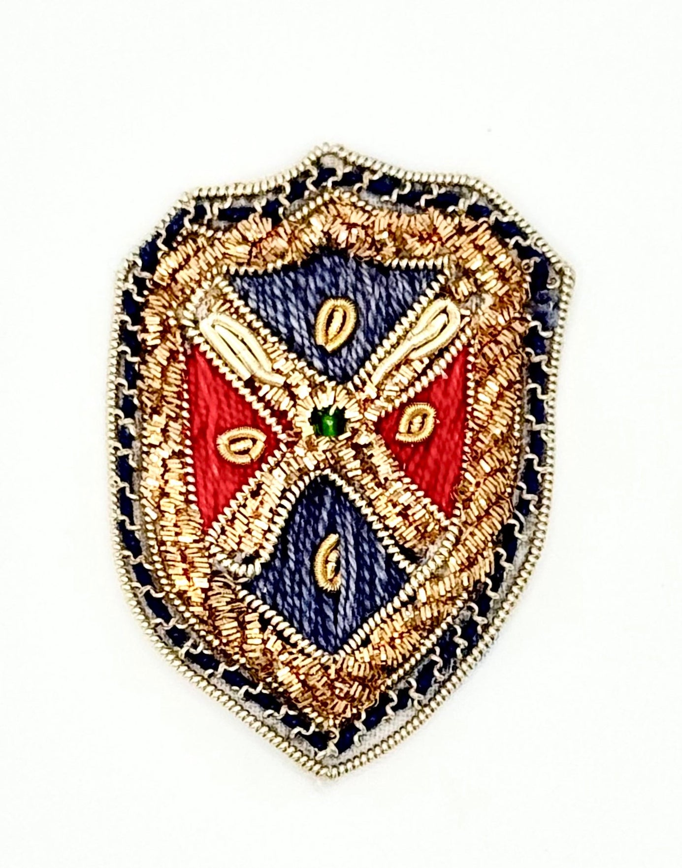 A shield-shaped embroidered patch featuring a detailed heraldic design with red and blue sections, gold and beige elements, a small green center jewel, and a embroidered border on a plain white background.