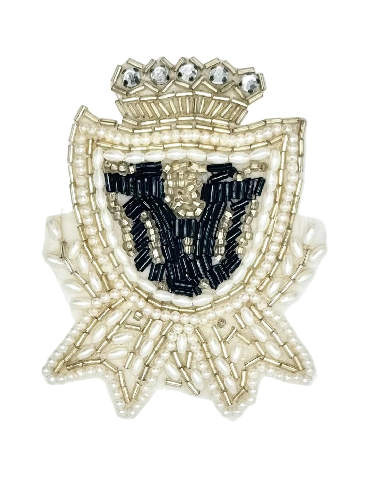 A decorative embroidered or beaded crest patch featuring a crown at the top, a shield shape outlined in pale gold and pearl-like beads, and a central black embroidered motif on a white background.