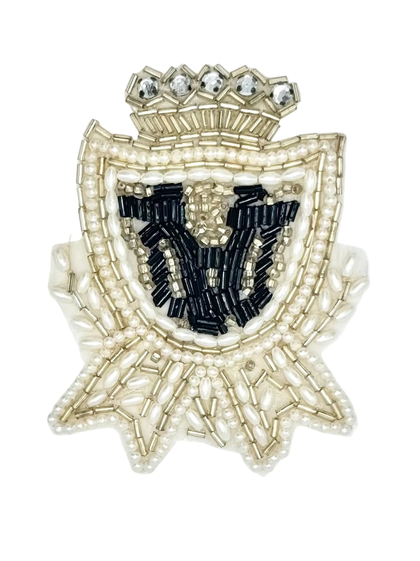 A decorative embroidered or beaded crest patch featuring a crown at the top, a shield shape outlined in pale gold and pearl-like beads, and a central black embroidered motif on a white background.