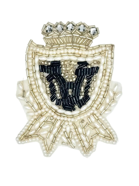 A decorative embroidered or beaded crest patch featuring a crown at the top, a shield shape outlined in pale gold and pearl-like beads, and a central black embroidered motif on a white background.