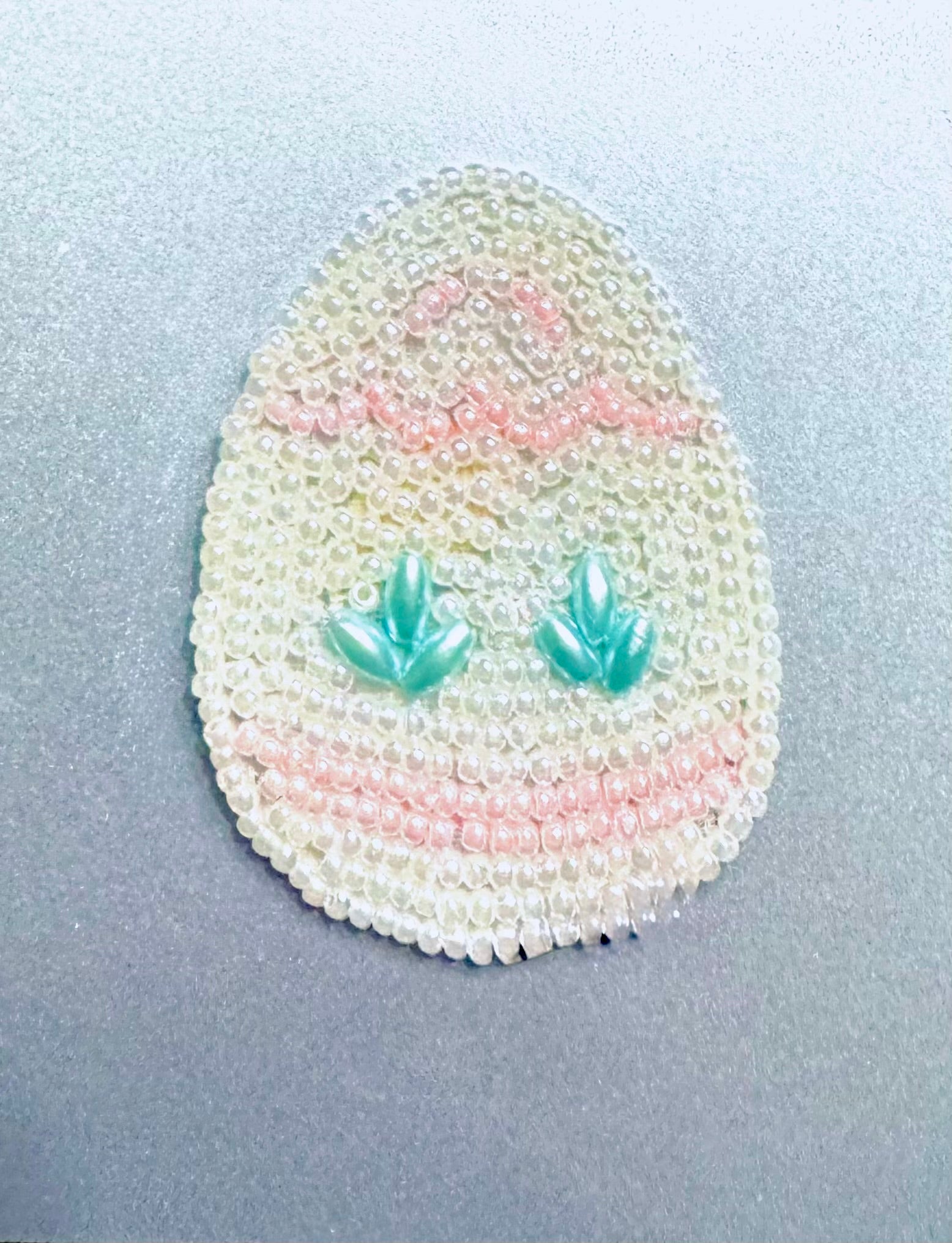 Egg-shaped beaded ornament with teal leaf designs on a light gray background