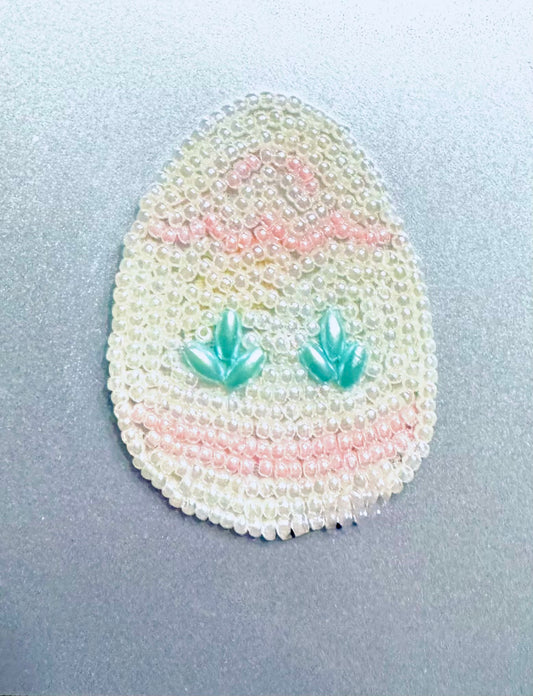 Egg-shaped beaded ornament with teal leaf designs on a light gray background