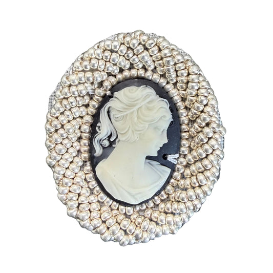 An oval vintage-style cameo brooch with a white profile portrait on a black background, surrounded by a textured silver beaded border, shown on a white surface.